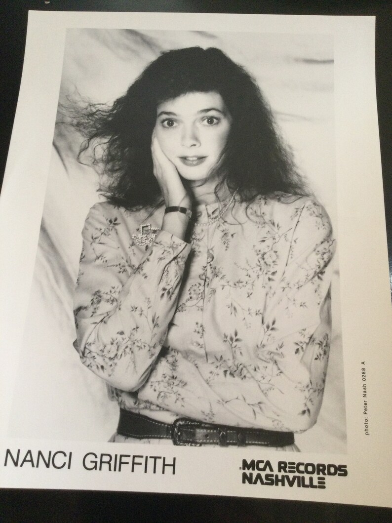 May include: Black and white photograph of Nanci Griffith, with long, dark, curly hair. She wears a floral print blouse, a belt, and a watch. The image includes the text "NANCI GRIFFITH" and "MCA RECORDS NASHVILLE."