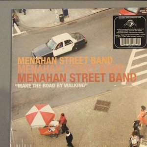 May include: Album cover for the Menahan Street Band's "Make the Road by Walking." The cover features an aerial view of a city street with a black and white car, a vendor cart, and pedestrians. The band's name is in orange text.