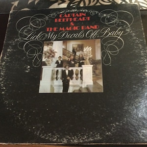 May include: Vintage vinyl record album cover for Captain Beefheart & The Magic Band's "Lick My Decals Off, Baby." The cover is black with the album title and band name in white and red script. A photo of the band is in the center.