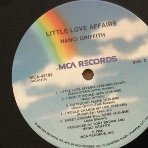 May include: A vinyl record for "Little Love Affairs" by Nanci Griffith. The record label is blue with a rainbow and clouds design. The text includes song titles and credits, with "MCA Records" branding.