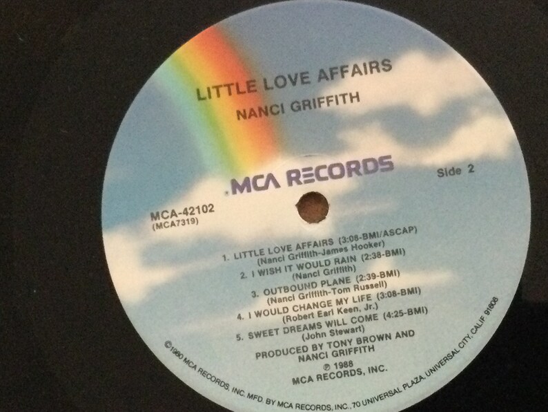 May include: A vinyl record for "Little Love Affairs" by Nanci Griffith. The record label is blue with a rainbow and clouds design. The text includes song titles and credits, with "MCA Records" branding.