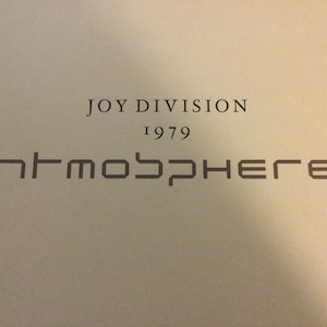 May include: A light-colored surface featuring the black text JOY DIVISION, the year 1979, and the word ATMOSPHERE written in a stylized, geometric font with some letters mirrored or inverted, creating a distinct graphic design layout.