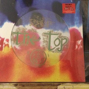 May include: The Cure's album "The Top" on vinyl. The album cover features a clear, circular overlay with the band's name and album title in green. The background has a colorful abstract design with red, orange, blue, and black hues.