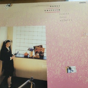 May include: Album cover for Nanci Griffith's "Little Love Affairs." The cover has a pink background with a repeating floral pattern. A photograph of a person in a black outfit is on the left. The album title and artist's name are printed in black.