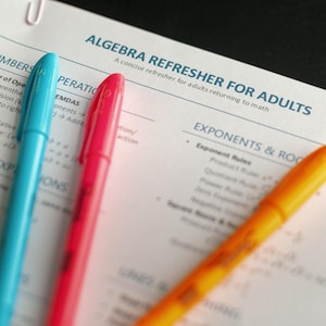 May include: Close-up of a worksheet titled "ALGEBRA REFRESHER FOR ADULTS" with blue, red, and orange pens. The worksheet covers topics like exponents and roots. A paper clip is visible at the top.
