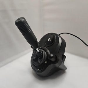 May include: Black Logitech G shifter for sim racing. The unit features a black gear shift knob, a leather-like boot, and a base with the Logitech G logo. A black cable extends from the unit.