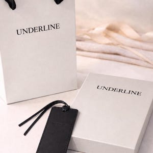 Può includere: Un sacchetto regalo in carta bianca con manici in nastro nero, una scatola regalo rettangolare bianca e un'etichetta rettangolare nera, tutti con la parola UNDERLINE stampata in lettere maiuscole nere sulle rispettive superfici.
