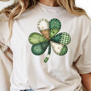 Può includere: T-shirt color crema con un grande design a trifoglio cucito. Il trifoglio è composto da varie tonalità di verde, con motivi a tinta unita e a scacchi, e dettagli di cucitura bianchi. Grafica a tema San Patrizio.
