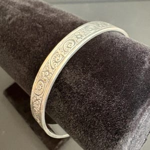 May include: A silver-toned metal bangle bracelet featuring a continuous embossed pattern of small flowers and swirling vine motifs. The surface of the bracelet has a polished finish with darkened recessed areas that highlight the intricate floral design.