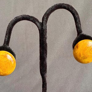 May include: A pair of round stud earrings featuring a polished, marbled amber-colored surface with swirling golden and brown patterns. The circular discs have a smooth, glossy finish and are displayed on a black velvet-textured earring stand.