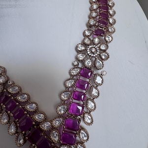 May include: A necklace featuring a series of rectangular deep purple stones arranged in a central column, flanked by teardrop-shaped clear stones and small round clear stones set in a metallic frame with a textured, antique-style finish.