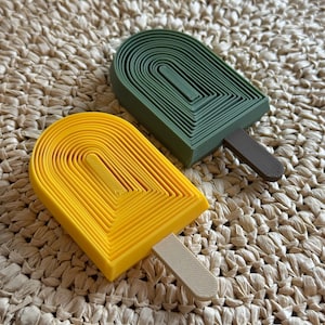 May include: Two popsicle-shaped objects, one yellow and one green, with brown and beige sticks. The popsicle shapes have a layered design. The objects are on a woven, beige surface.