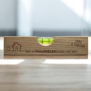 Könnte beinhalten: Ein Holz-Wasserwaage mit einer grünen Libelle. Auf der Wasserwaage befinden sich eine Hausgrafik, der Text "Damit der Haussegen niemals schief hängt" und die Namen "Laura & Sebastian".