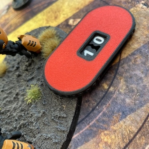 Könnte beinhalten: Ein roter, rechteckiger Spielzähler mit schwarzem Rand und einem kleinen Fenster mit der Zahl 10. Der Zähler ist strukturiert und scheint aus Kunststoff zu sein. Das Bild stammt aus einer Tabletop-Gaming-Umgebung.