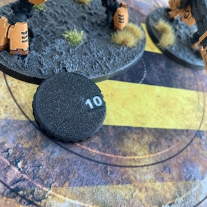 Könnte beinhalten: Ein runder schwarzer Spielmarker aus Kunststoff mit strukturierter Oberfläche und der weißen Zahl 10, die auf einer kleinen vertieften Lasche aufgedruckt ist. Der Marker dient zur Verfolgung von Spielstatistiken und liegt auf einer flachen Oberfläche.