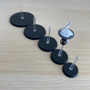 Könnte beinhalten: Eine Ansammlung schwarzer, kreisförmiger Sockel mit transparenten Kunststoffstielen, die zum Halten von Miniaturfiguren bestimmt sind. Ein Sockel trägt eine detaillierte Miniatur mit grauen und schwarzen Komponenten. Die Sockel sind auf einer hellen Holzmaserung angeordnet.