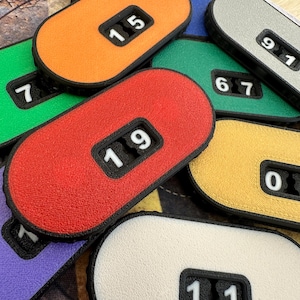May include: A collection of oval-shaped plastic counters in various colours including red, orange, yellow, green, purple, and white. Each counter features a black rectangular window displaying two white numbers, such as 19, 15, 91, 67, 0, and 11.