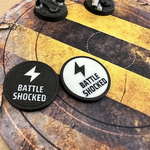 以下が含まれることがあります： 稲妻のアイコンと「BATTLE SHOCKED」という文字が刻まれた2つの円形ゲームトークン。1つは黒い背景に白の文字とアイコン、もう1つは白い背景に黒の文字とアイコンが施されています。どちらも黒い縁取りがされています。