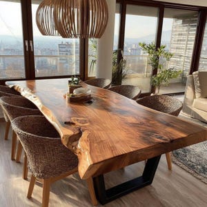 May include: A large, natural-edge wooden dining table with a dark finish, supported by a black metal base. Woven wicker chairs surround the table. Two pendant lights hang above, and a city view is visible through the windows.