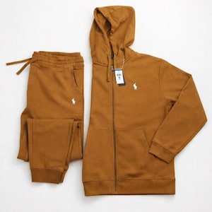 May include: A brown hooded zip-up sweatshirt and matching sweatpants set. The sweatshirt has a white logo on the chest and a drawstring hood. The sweatpants have an elastic waistband and a white logo. The set is made of a soft, comfortable material.