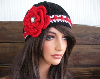 May include: A black crocheted beanie featuring a large, layered red yarn flower on the side. The flower has a central white pearl-like accent. A horizontal band of red and white stitching encircles the base of the black knit hat.