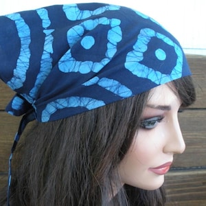 May include: A dark navy blue head scarf featuring a repeating light blue tie-dye pattern of circular and square shapes. The fabric is tied at the back with two thin fabric strips, creating a secure fit around the head with a visible knotted tail.