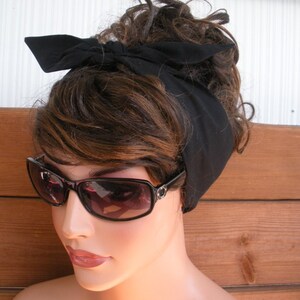 May include: A black fabric headband tied in a bow on top of a woman's head. The headband is worn over brown hair and the woman is wearing sunglasses.