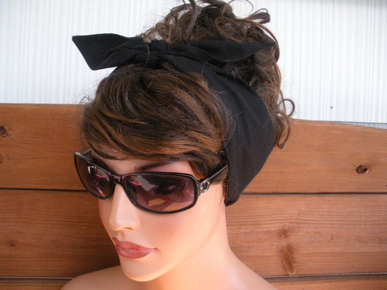 May include: A black fabric headband tied in a bow on top of a woman's head. The headband is worn over brown hair and the woman is wearing sunglasses.