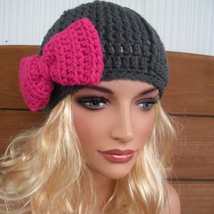 May include: A dark gray crocheted beanie hat with a large, bright pink bow on the side. The hat is handmade and appears to be soft and warm, suitable for cooler weather. The bow adds a decorative touch.