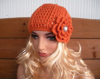 May include: An orange crochet hat with a floral embellishment. The hat is a warm, burnt orange color and features a large, crocheted flower on the side, adorned with a pearl-like center. The hat is a stylish accessory.