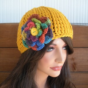 Op de afbeelding: Mosterdgele gehaakte hoed met een grote, veelkleurige bloemenapplicatie. De bloem heeft tinten rood, groen, blauw en paars, met een gele knoop in het midden. De hoed is een beanie-stijl, geschikt voor koeler weer.