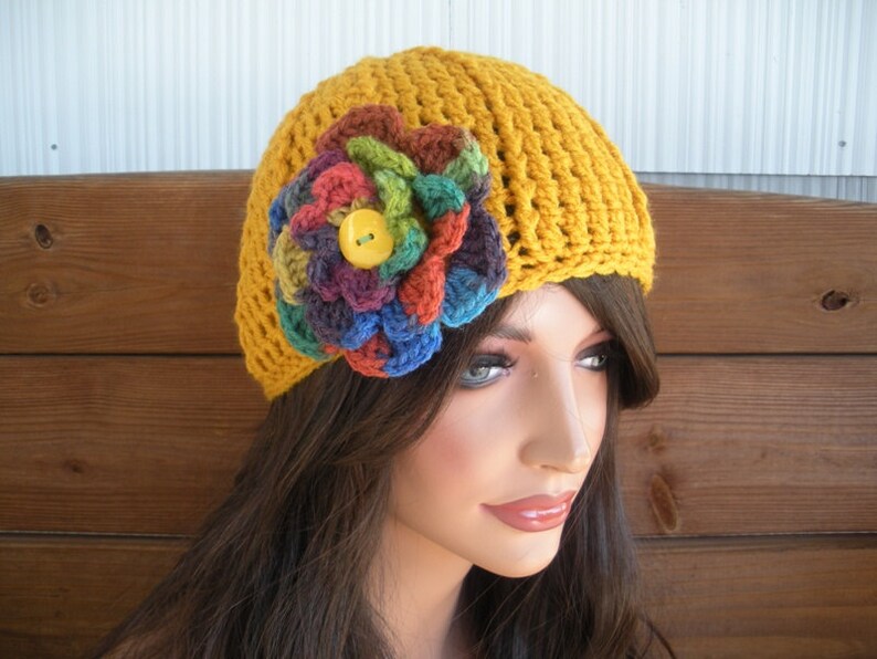 Op de afbeelding: Mosterdgele gehaakte hoed met een grote, veelkleurige bloemenapplicatie. De bloem heeft tinten rood, groen, blauw en paars, met een gele knoop in het midden. De hoed is een beanie-stijl, geschikt voor koeler weer.