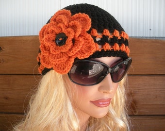 May include: A black and orange crocheted beanie with a large orange flower on the side. The beanie has a crocheted design with a black button in the center of the flower.