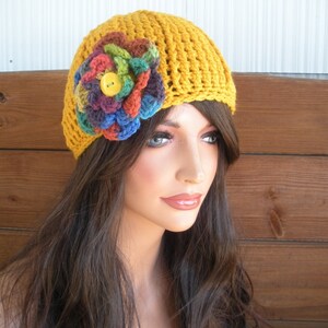 Op de afbeelding: Mosterdgele gehaakte hoed met een grote, kleurrijke bloemenapplicatie. De bloem heeft tinten blauw, groen, rood en paars, met een gele knoop in het midden. De hoed wordt gedragen door een mannequin met lang bruin haar.