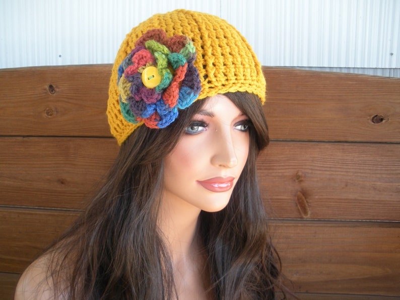 Op de afbeelding: Mosterdgele gehaakte hoed met een grote, kleurrijke bloemenapplicatie. De bloem heeft tinten blauw, groen, rood en paars, met een gele knoop in het midden. De hoed wordt gedragen door een mannequin met lang bruin haar.