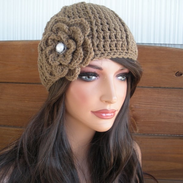 May include: A brown crochet beanie featuring a textured knit pattern and a large, layered crochet flower accent on the side. The centre of the flower is adorned with a round, white faux pearl cabochon set in a silver-toned metal frame.