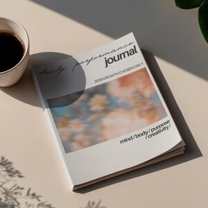 Puede incluir: Un diario blanco con las palabras "daily performance journal" y "2026 GROWTH CHOSEN DAILY" impresas en la portada. El diario también incluye las palabras "mind / body / purpose / creativity". Una taza de café está cerca.