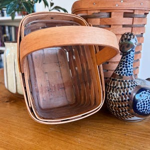 May include: A wooden basket with a hinged lid and a curved handle. The basket is made of light brown wood and has a rectangular shape. A ceramic duck figurine with blue and brown patterns sits nearby. A second basket is in the background.