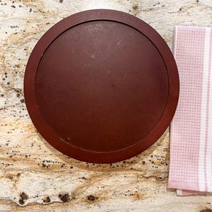 Puede incluir: Una bandeja redonda de madera marrón oscuro con un borde elevado. La bandeja está colocada sobre una superficie moteada de color marrón claro, con una toalla a rayas rosas y blancas en la esquina. La bandeja probablemente se utiliza para servir o exhibir.