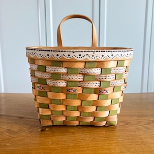 May include: Woven basket with a wooden handle. The basket features a light brown and green woven pattern, accented with white lace and small floral designs. The top edge is decorated with a scalloped design.