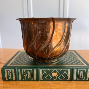 May include: A copper-toned metal planter with a scalloped rim and a swirling ribbed texture, resting on a dark green hardcover book titled Great Cases of Sherlock Holmes by Sir Arthur Conan Doyle, featuring gold-colored decorative grid patterns.