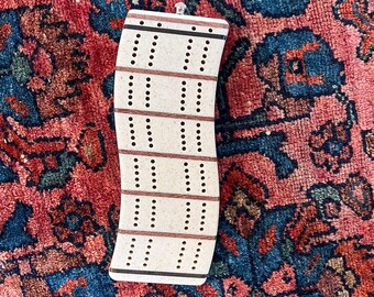 May include: A cream-coloured cribbage board with brown and black accents. The board has multiple rows of holes for pegs, with a curved design. A small metal loop is at the top. The board is on a patterned rug.