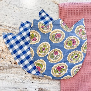 May include: Three tulip-shaped quilted pot holders. One is blue and white gingham, and the other two are light blue with floral patterns in pink, green, and yellow. A pink waffle-weave towel is on the right.
