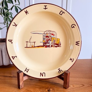 May include: A cream-colored ceramic plate with a dark brown rim featuring a central illustration of a covered wagon and a person cooking over a fire. The border is decorated with dark brown cattle brand symbols arranged in a circular pattern.