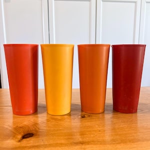 May include: Four tall, cylindrical plastic tumblers arranged in a row. The cups feature a smooth, matte finish and are colored in a gradient of warm tones, specifically bright orange, golden yellow, burnt orange, and deep red, standing upright.