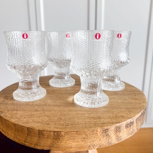 May include: Four clear textured glass goblets with a red "i" sticker on each, arranged on a round wooden table. The goblets have a unique bark-like texture and a flared base.