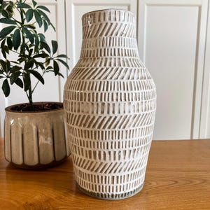 May include: A tall, off-white ceramic vase with a textured, geometric pattern. The vase has a wide base that tapers to a narrower neck and a flared rim. The design features vertical and diagonal lines. A potted plant is in the background.