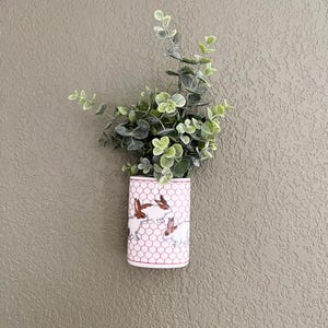 Könnte beinhalten: Eine weiße und rosa Keramik-Wandvase mit Hasenmuster, gefüllt mit grünem und grauem künstlichem Eukalyptus. Die Vase hat ein sechseckiges Muster und ist an einer strukturierten, neutral getönten Wand befestigt. Die Vase ist etwa 15 cm hoch.