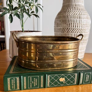 May include: A vintage brass planter with two handles, resting on a green book with gold lettering. A white vase with a textured pattern and a potted plant are in the background. The planter is oval-shaped.