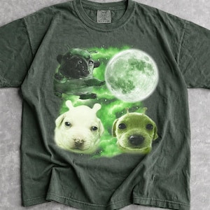 Könnte beinhalten: Dunkelgrünes T-Shirt mit einer Grafik von drei Welpen und einer Möpse, jeweils mit außerirdischem Aussehen, vor einem grünen Weltraumhintergrund mit einem großen grünen Mond. Das T-Shirt hat einen Rundhalsausschnitt und kurze Ärmel.
